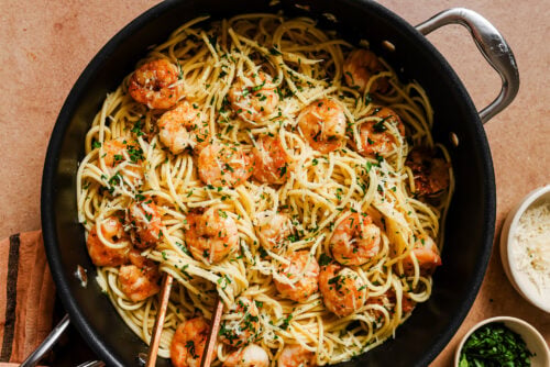 A skillet filled with Garlic Shrimp Spaghetti, sautéed shrimp, chopped parsley, and grated cheese, with tongs resting on top. Small bowls of cheese and herbs are nearby on a light brown surface.