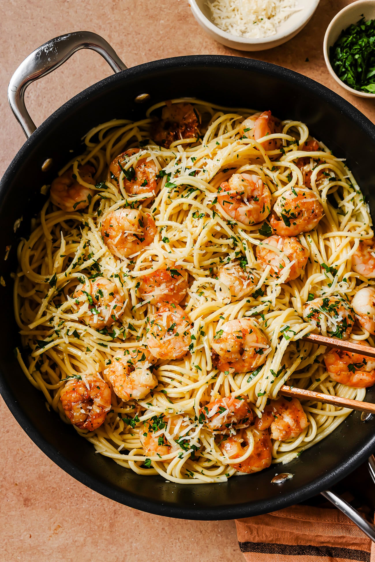 A skillet filled with Garlic Shrimp Spaghetti, sautéed shrimp, chopped parsley, and grated cheese, with tongs resting on top. Small bowls of cheese and herbs are nearby on a light brown surface.
