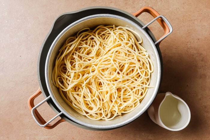 A pot filled with cooked Garlic Shrimp Spaghetti sits on a beige surface. Next to it is a small white pitcher containing pasta water.