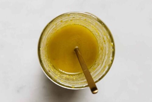A glass filled with yellow salad dressing, perfect for a Breakfast Salad Recipe, with a gold spoon resting inside, viewed from above on a white surface.
