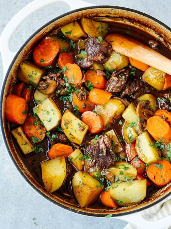 A pot of Dutch Oven Beef Stew, filled with chunks of beef, potatoes, carrots, and mushrooms simmered in a rich brown broth and garnished with parsley. A wooden spoon rests inside the hearty stew.
