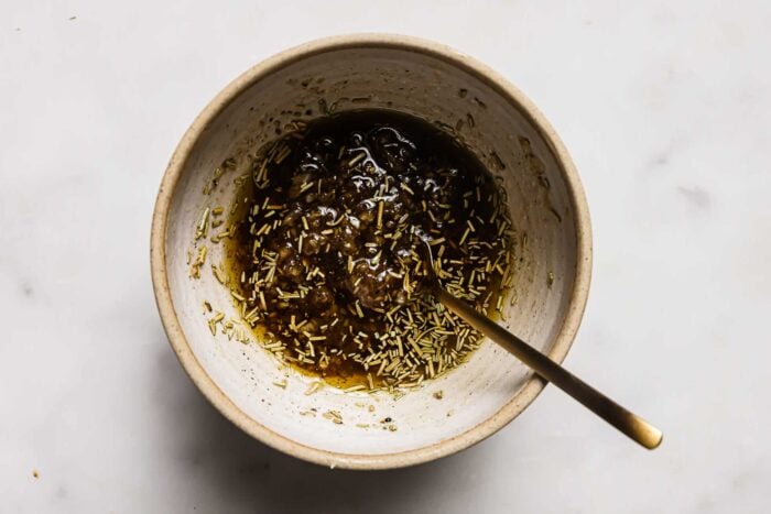 A white ceramic bowl with a brown liquid marinade for Bacon Wrapped Pork Tenderloin, mixed with herbs, and a spoon resting inside, on a light marble surface.