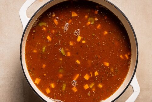 A pot filled with Pasta e Fagioli Soup, a tomato-based soup featuring diced carrots, celery, herbs, and beans, seen from above on a light-colored surface.