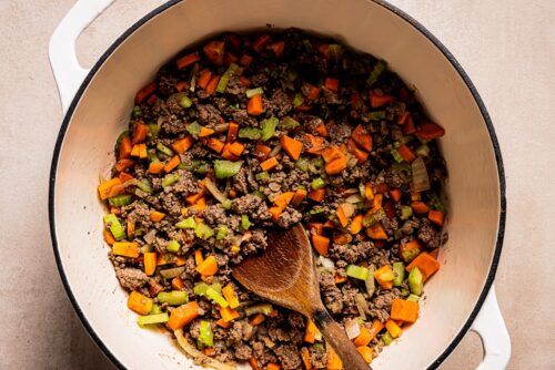 A white pot filled with cooked ground beef, diced carrots, green bell peppers, and onions is being stirred with a wooden spoon—perfect base ingredients for a hearty Pasta e Fagioli Soup.