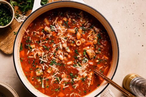 A pot of hearty Pasta e Fagioli Soup with beans, pasta, vegetables, tomato broth, and topped with grated cheese and parsley sits ready to serve. Fresh herbs and extra cheese rest nearby on a wooden board.