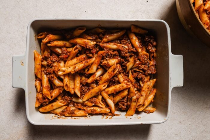 A white baking dish filled with Baked Ziti with Ground Beef and tomato sauce sits on a light-colored surface. A partial view of another bowl of pasta is visible in the top left corner.