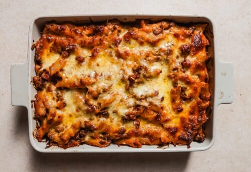 A rectangular baking dish filled with Baked Ziti with Ground Beef, topped with melted, golden-brown cheese and a tomato-based sauce, sits on a light-colored surface.