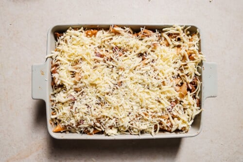 A rectangular baking dish filled with Baked Ziti with Ground Beef is topped with a generous, even layer of shredded cheese, ready to be baked. The dish rests on a light-colored countertop.