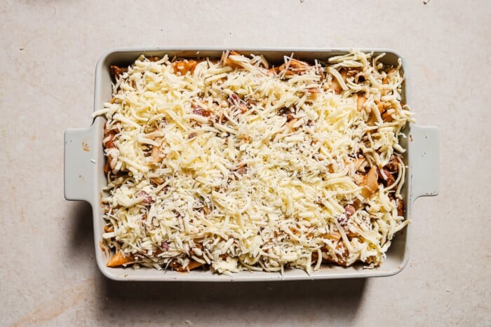 A rectangular baking dish filled with ziti is topped with a generous, even layer of shredded cheese, ready to be baked. The dish rests on a light-colored countertop.