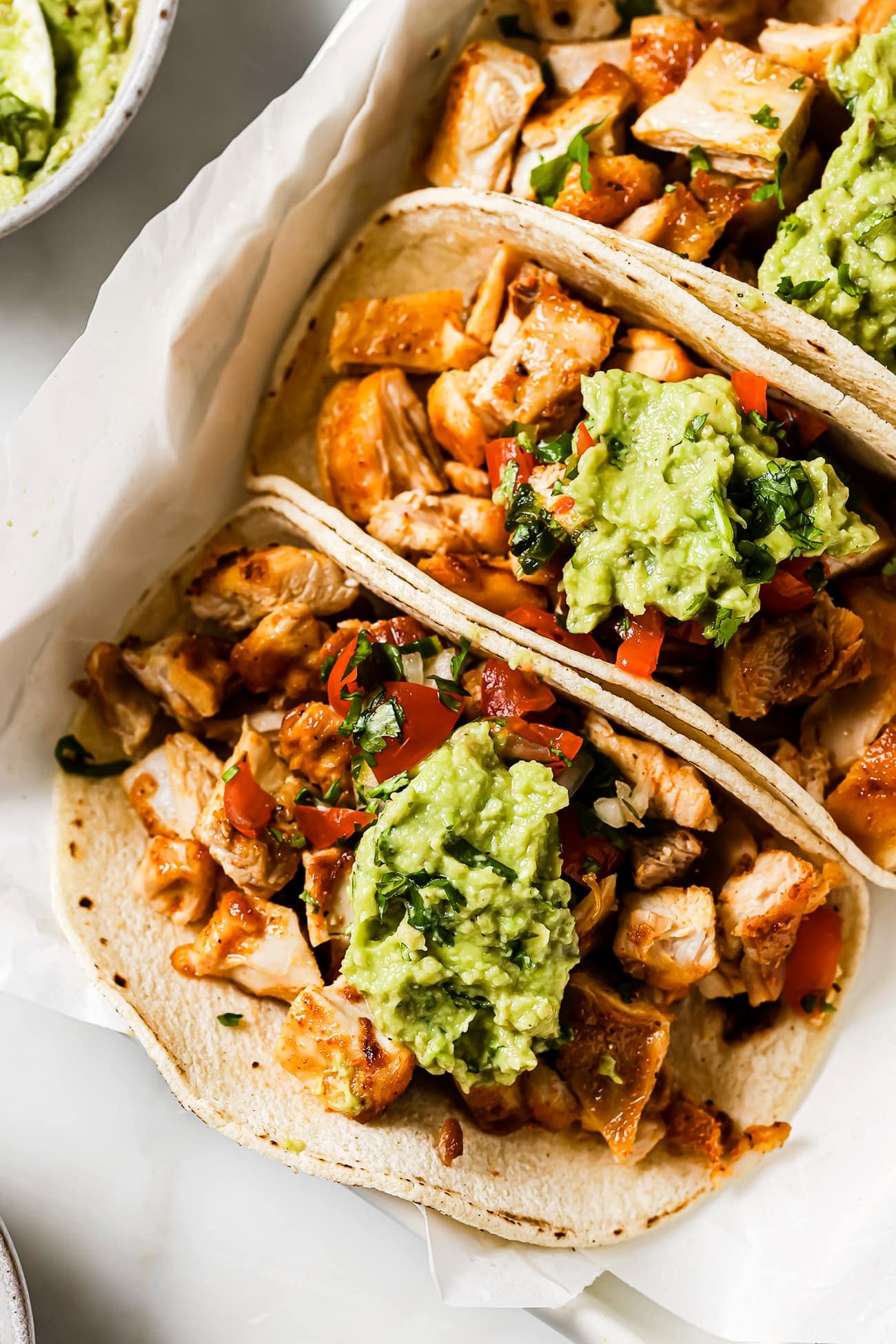 Three chicken tacos topped with Pico de Gallo and guacamole on parchment paper.