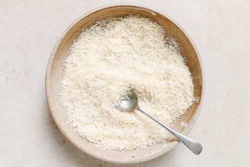 A bowl filled with finely grated parmesan cheese, perfect for topping Air Fryer Chicken Cutlets, with a metal spoon resting inside, placed on a light-colored surface.