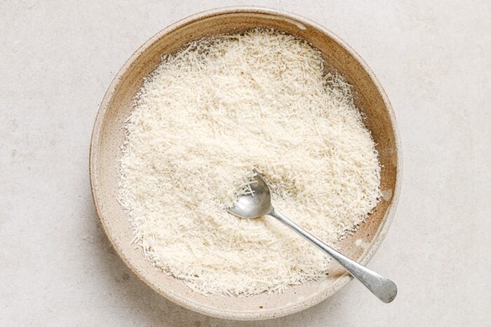 A bowl filled with finely grated parmesan cheese, perfect for topping Air Fryer Chicken Cutlets, with a metal spoon resting inside, placed on a light-colored surface.