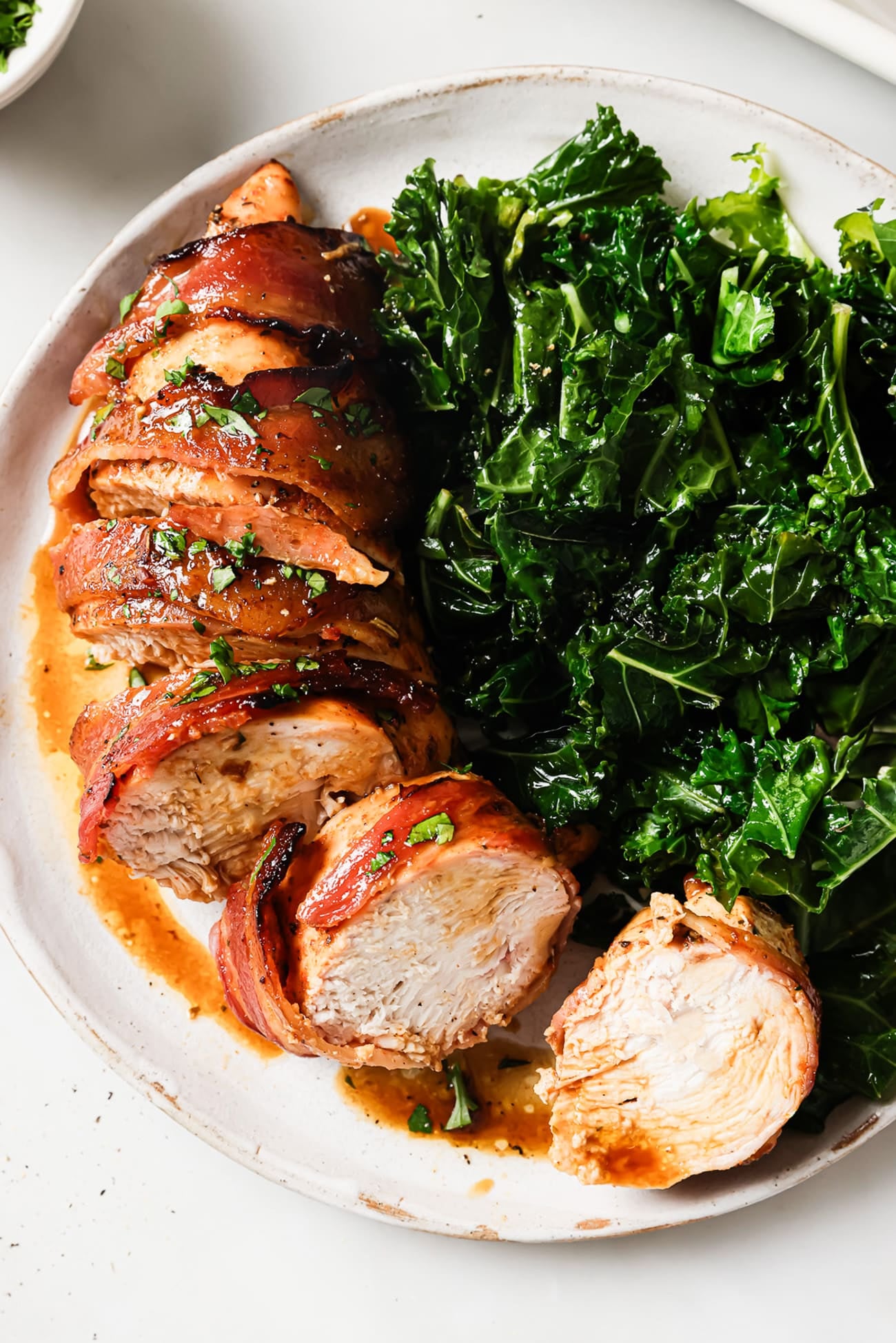 A sliced bacon-wrapped chicken breast next to a side of wilted kale on a white plate. 