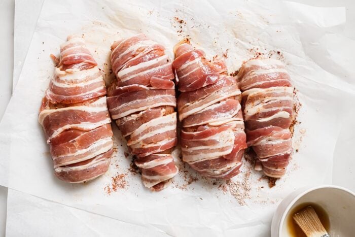 Four Bacon Wrapped Chicken Breasts sit on white parchment paper, sprinkled with seasoning. A small bowl with a brush is visible in the lower right corner of the image, ready for basting.