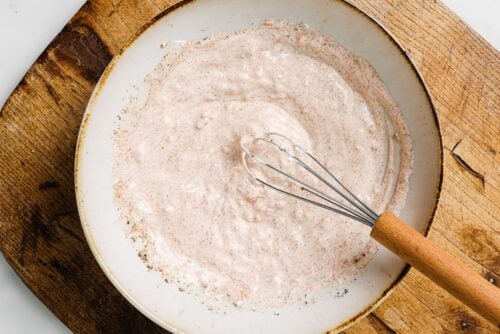 A bowl of creamy, light pink sauce—perfect for Yogurt Marinated Chicken Breast—speckled with seasoning is whisked with a metal whisk with a wooden handle, set on a wooden surface.