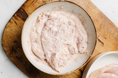 Yogurt Marinated Chicken Breast pieces coated in a creamy, seasoned marinade sit in a shallow white bowl on a wooden cutting board, with a plate of plain chicken breasts nearby.