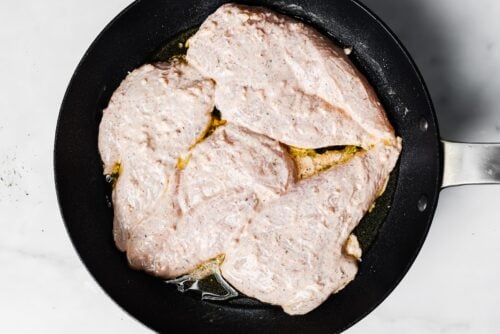 Raw, seasoned Yogurt Marinated Chicken Breasts are coated in a creamy marinade and spread out in a black frying pan with some oil, ready to be cooked, photographed from above on a white surface.