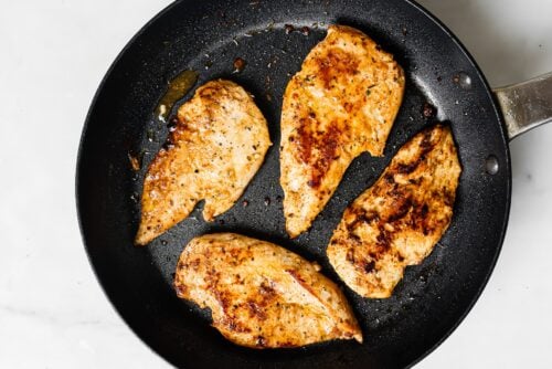 Four golden brown Yogurt Marinated Chicken Breasts in a black skillet on a white surface. The chicken fillets show visible spices and searing marks, indicating they're perfectly seasoned and cooked.