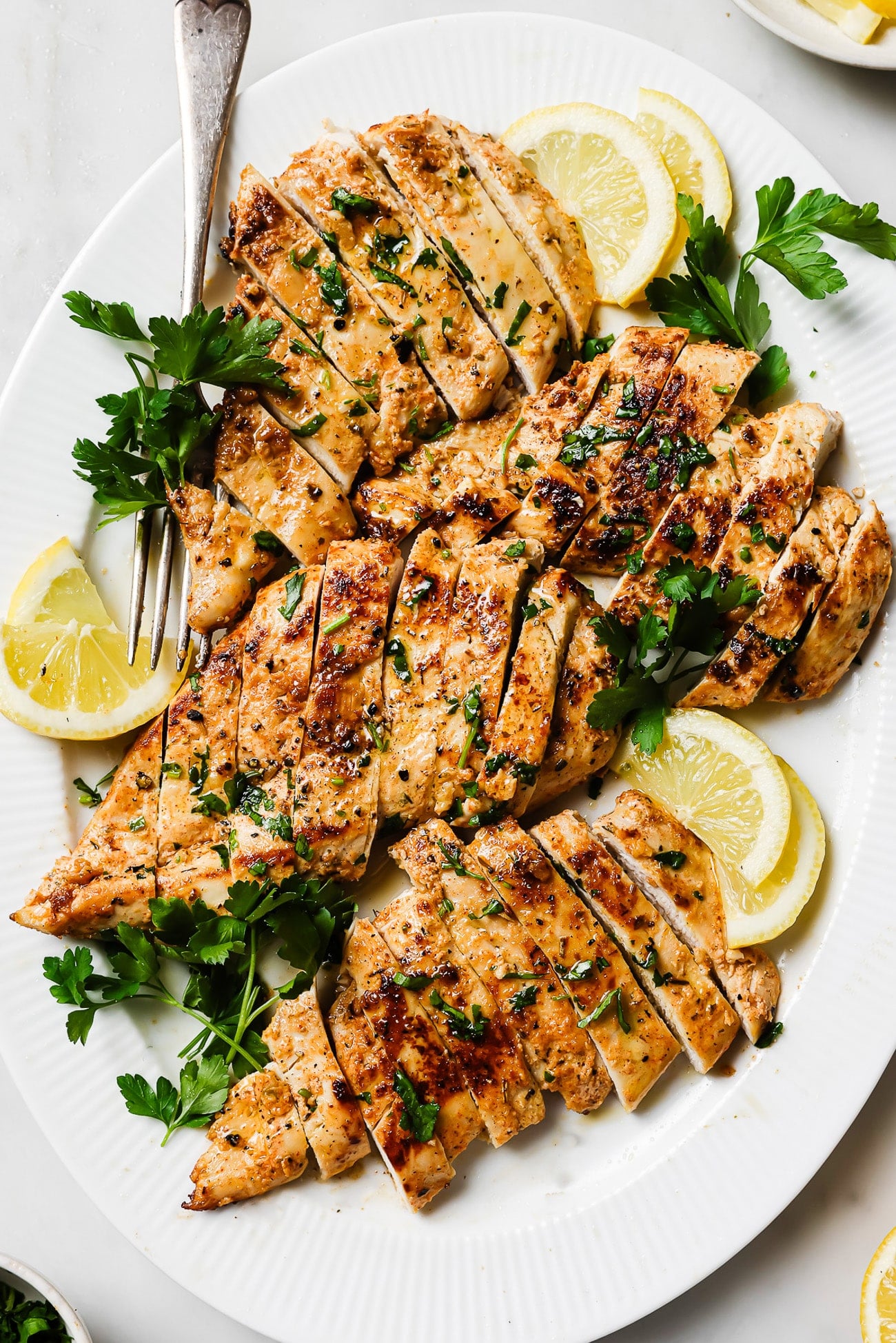 Yogurt-marinated chicken breast sliced and garnished with parsley and lemon wedges on a white serving plate. 