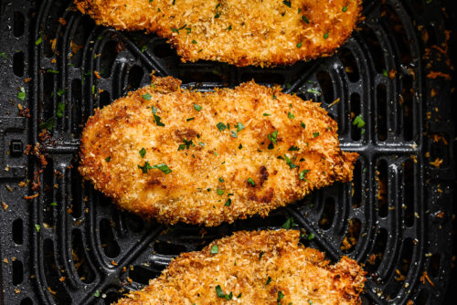 Three golden Air Fryer Chicken Cutlets rest in a basket, garnished with chopped parsley. The chicken breast looks crispy and perfectly cooked for a delicious meal.
