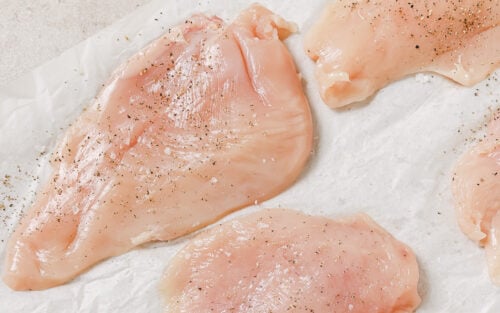 Raw chicken breasts seasoned with salt and pepper are arranged on a sheet of white parchment paper, ready to be transformed into crispy Air Fryer Chicken Cutlets on a light-colored surface.