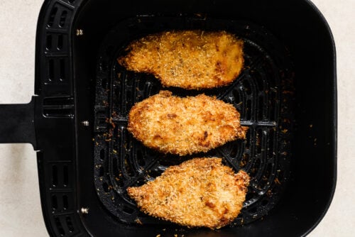 Three Air Fryer Chicken Cutlets are cooking inside a black air fryer basket, each with a golden-brown crispy coating. The vented, ridged bottom ensures even cooking for these delicious cutlets.