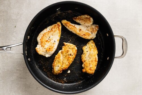 Four pieces of seasoned, golden-brown chicken breasts in a black skillet on a light gray surface—perfect for making Creamy Mushroom Chicken.