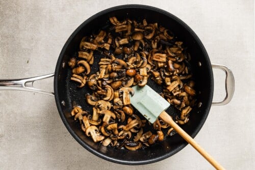 A black skillet filled with sautéed mushrooms for creamy mushroom chicken, stirred with a light green spatula on a light-colored countertop.