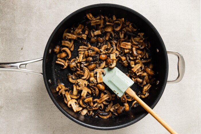 A black skillet filled with sautéed mushrooms for creamy mushroom chicken, stirred with a light green spatula on a light-colored countertop.