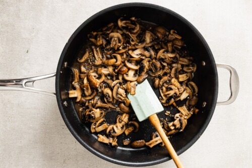A black skillet filled with sautéed sliced mushrooms sits on a light-colored surface, hinting at the beginnings of a creamy mushroom chicken dish. A green spatula with a wooden handle rests inside the pan, stirring the mushrooms.
