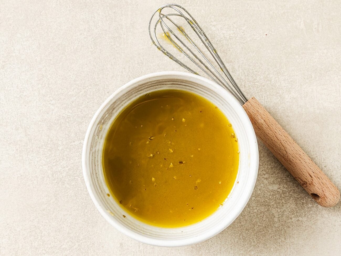 A wooden-handled whisk sits on the countertop next to a bowl of vinaigrette salad dressing.