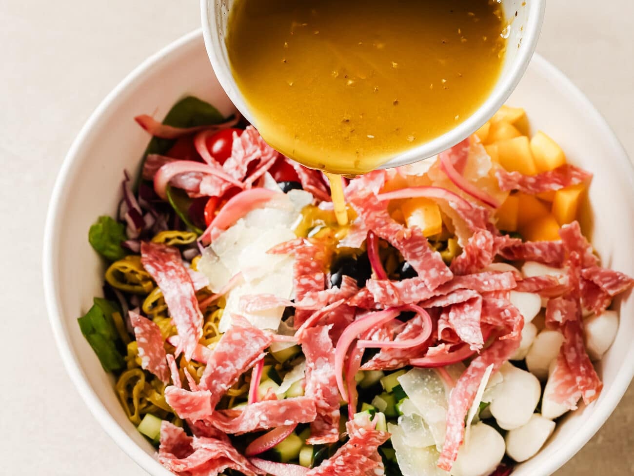 Pouring salad dressing from a small bowl onto the Italian chopped salad in a large serving bowl.