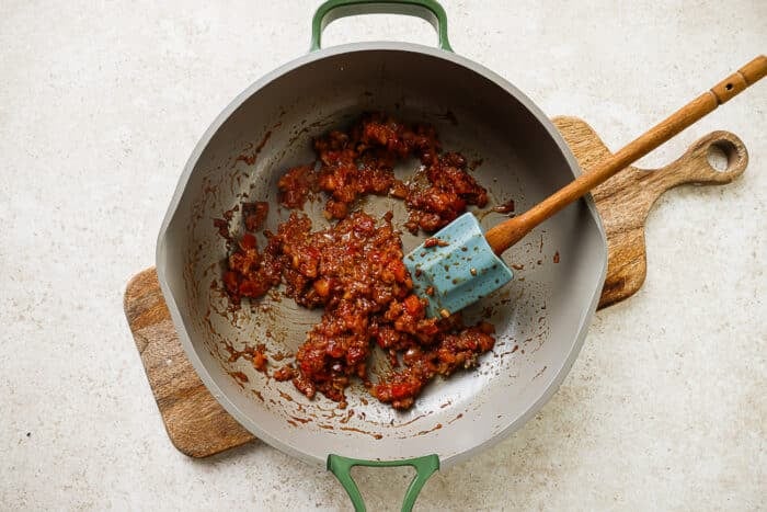 Tomato, onion, and spice paste cooking in a skillet with spatula on a wooden board.