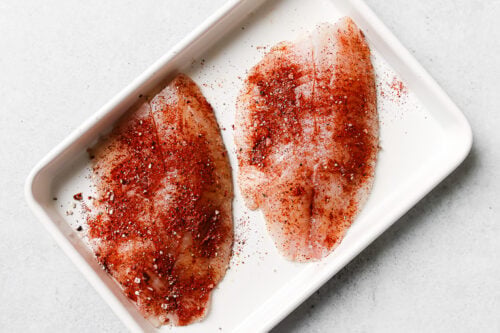 Seasoned raw tilapia fillet on a white baking sheet.