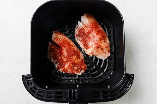 Raw tilapia fillets, seasoned and placed in an air fryer basket.