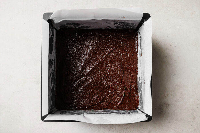 Chocolate brownie batter evenly spread in a parchment-lined square baking pan, ready to bake.