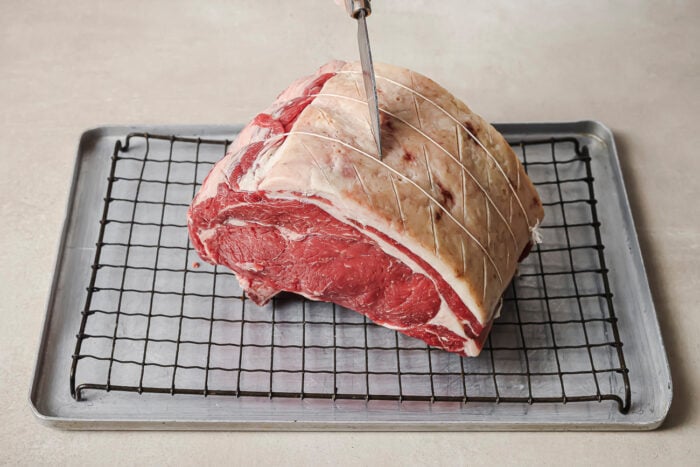 Scoring the fat cap of a raw prime rib roast on a wire rack to prepare it for reverse searing.