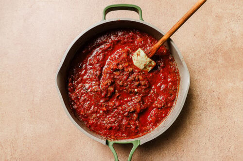 A green-handled pot filled with chunky red tomato sauce for Baked Rigatoni with Ground Beef sits on a beige surface. A wooden spatula rests in the thick, hearty sauce, rich with visible tomato and herb pieces.