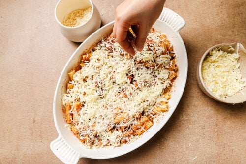 A hand sprinkling mozzarella cheese on top of the rigatoni with ground beef in a casserole dish.