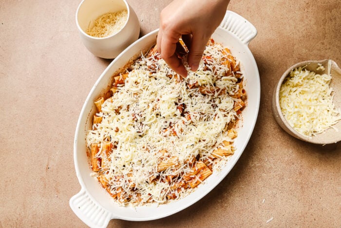 A hand sprinkling mozzarella cheese on top of the rigatoni with ground beef in a casserole dish.