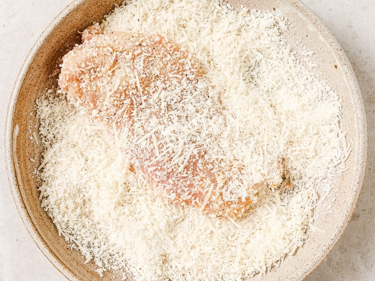 A raw breaded chicken breast, perfect for making Air Fryer Chicken Cutlets, is coated in shredded parmesan cheese and breadcrumbs, resting in a beige ceramic bowl on a light countertop.