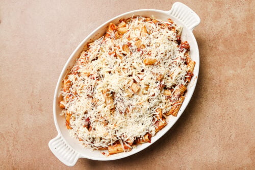 A white oval baking dish filled with Baked Rigatoni with Ground Beef, mixed with tomato sauce and topped with a generous layer of shredded cheese, ready to be baked. The dish sits on a light brown surface.
