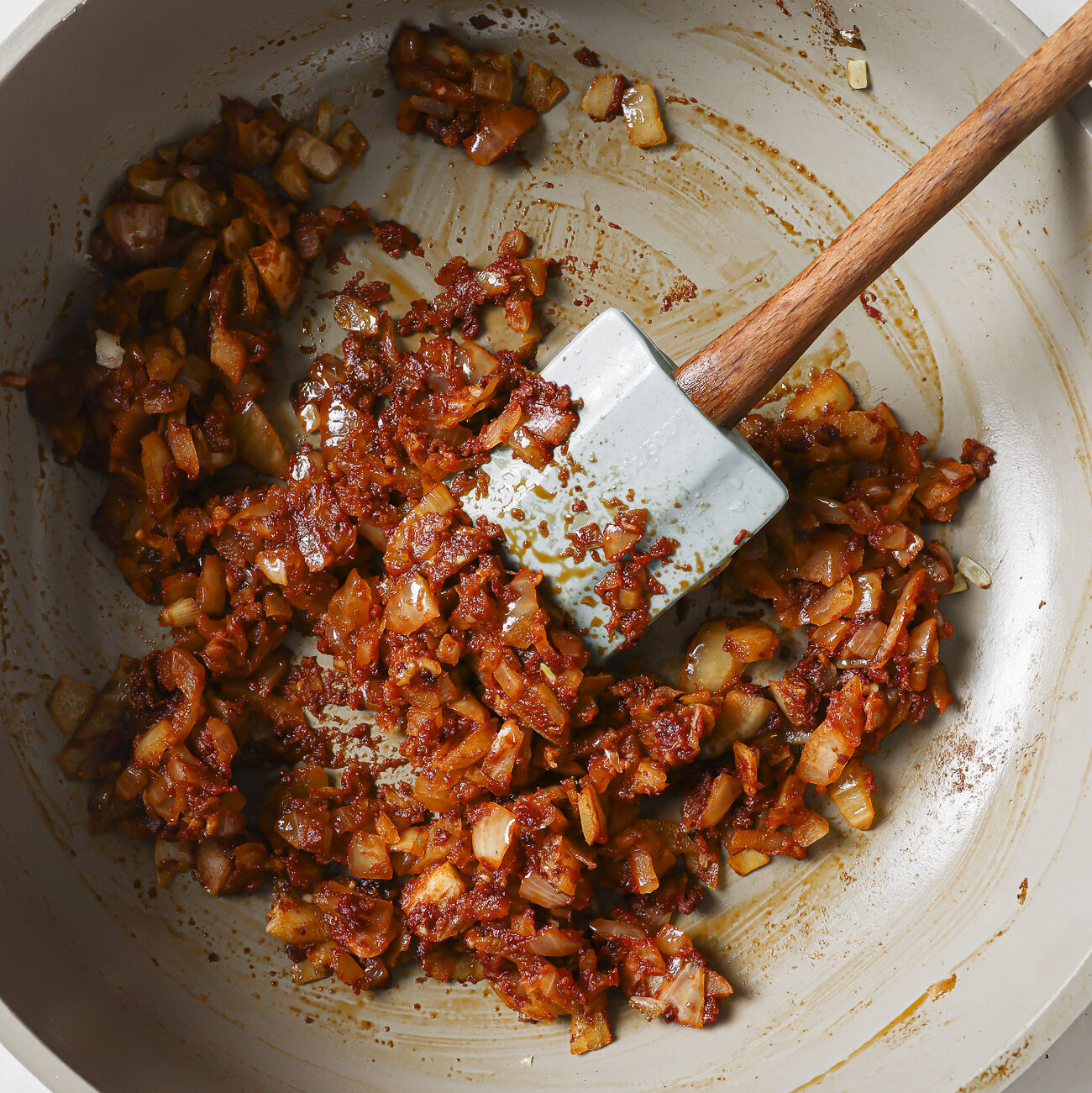 Chopped onions and spices are being sautéed in a large beige pan, stirred with a blue spatula—a perfect start for your Slow Cooker Chili Recipe. The mixture appears golden brown and well-cooked.