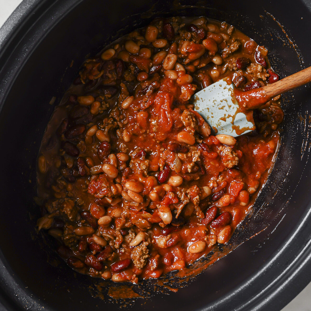 A slow cooker filled with a hearty Slow Cooker Chili Recipe—made of ground beef, kidney beans, white beans, and tomato sauce—being stirred with a blue spatula.
