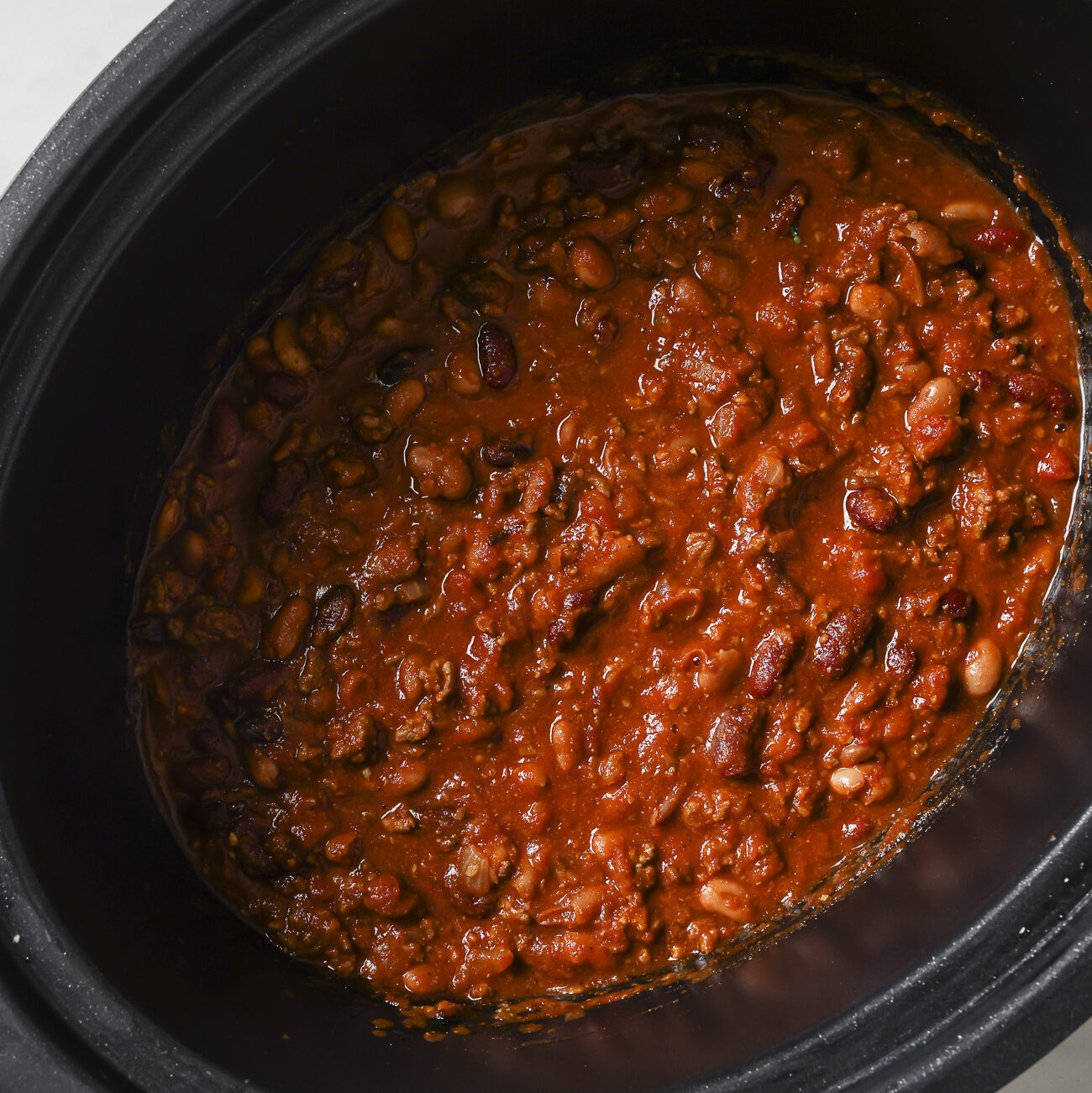 A slow cooker filled with thick, hearty Slow Cooker Chili Recipe made of beans, tomatoes, and ground meat sits on a white surface.