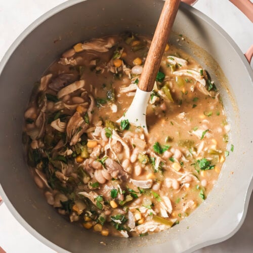 A large pot filled with creamy White Chicken Chili is being stirred with a white and wooden spoon. The soup features shredded chicken, corn, white beans, and greens on a light countertop.